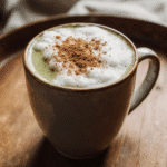 Cinnamon Matcha Latte – Cozy, Creamy, And Full Of Flavor! 8 A cozy ceramic mug filled with a creamy cinnamon matcha latte, topped with a swirl of matcha foam and cinnamon, sitting on a rustic wooden surface with warm natural lighting