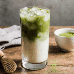 Iced Matcha Latte – Easy, Fresh & Delicious 11 Aesthetic iced matcha latte in a clear glass, creamy green tones with swirls of milk, styled on a wooden tray with matcha powder and bamboo whisk nearby